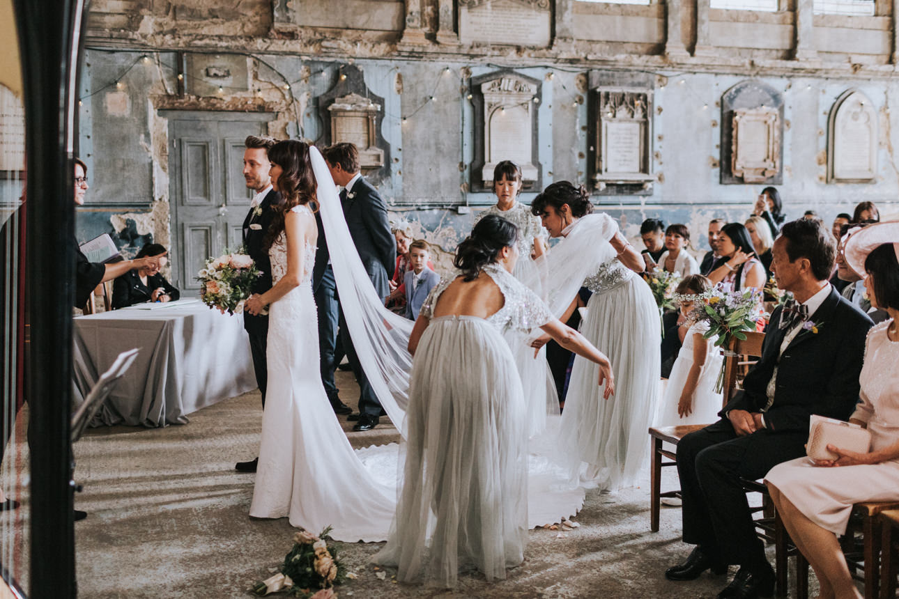 Bride at Asylum Chapel Wedding, her long veil being adjusted by bridesmaids in grey gowns. Guests seated, historic venue with faded walls.