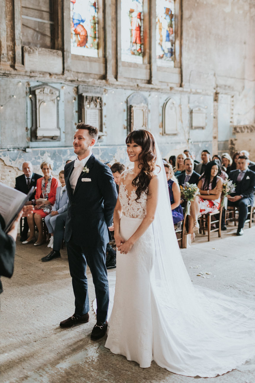 Bride and groom stand at the altar in a unique ceremony space with stained glass windows and aged walls. Guests are seated in the background.