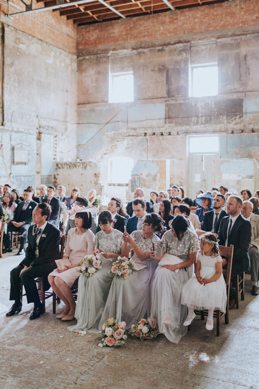 Guests Wedding watch the ceremony in a rustic, high-ceilinged space. Bridesmaids in silver gowns sit with bouquets; a flower girl in white completes the scene.